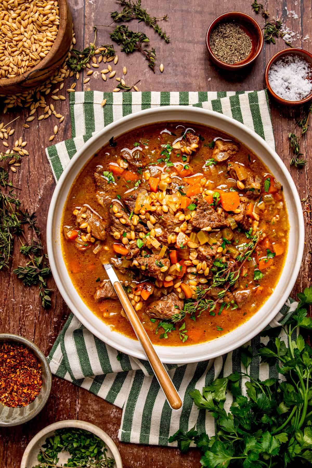 A bowl of beef and barley soup topped with fresh herbs sits on a striped towel, surrounded by spices, fresh thyme, a bowl of dry barley, and chopped parsley on a rustic wooden table.