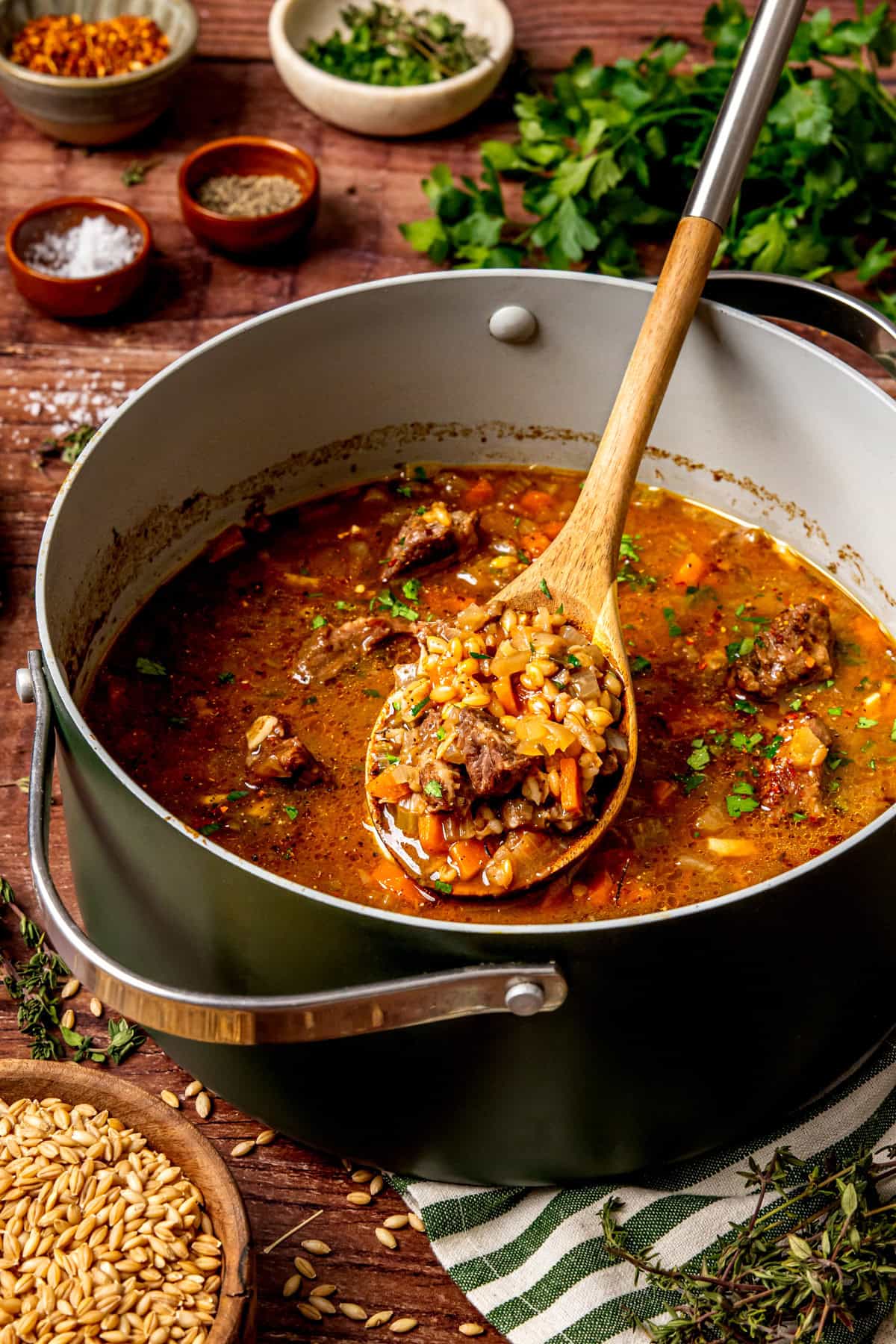 A large pot of beef and barley soup with chunks of beef, barley, carrots, and herbs. A wooden spoon holds a serving of the soup above the pot. Fresh parsley and small bowls of spices are on the wooden table nearby.