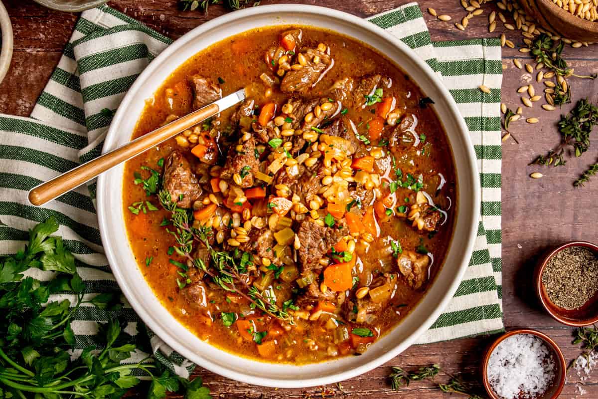 A bowl of hearty beef and barley soup with chunks of beef, carrots, and herbs, garnished with fresh parsley. The bowl is placed on a green-striped cloth with spices and herbs scattered around.