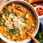 A bowl of hearty vegetable and lentil soup topped with shaved parmesan, fresh herbs, and chili flakes, with a wooden spoon, bread, and a small dish of chili flakes on the side.
