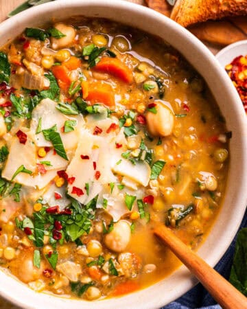 A bowl of hearty vegetable and lentil soup topped with shaved parmesan, fresh herbs, and chili flakes, with a wooden spoon, bread, and a small dish of chili flakes on the side.