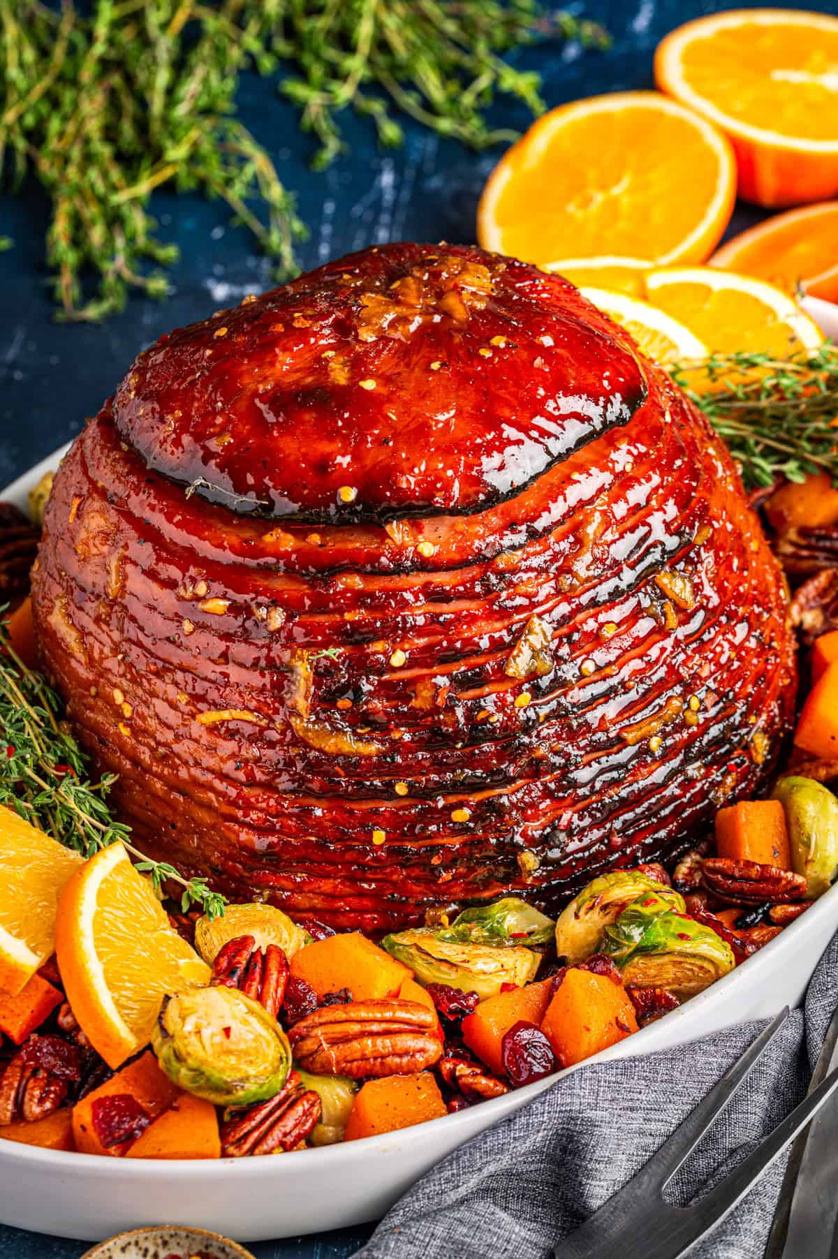 A glazed spiral ham garnished with orange slices, fresh herbs, pecans, brussels sprouts, and roasted squash, arranged on a white platter.