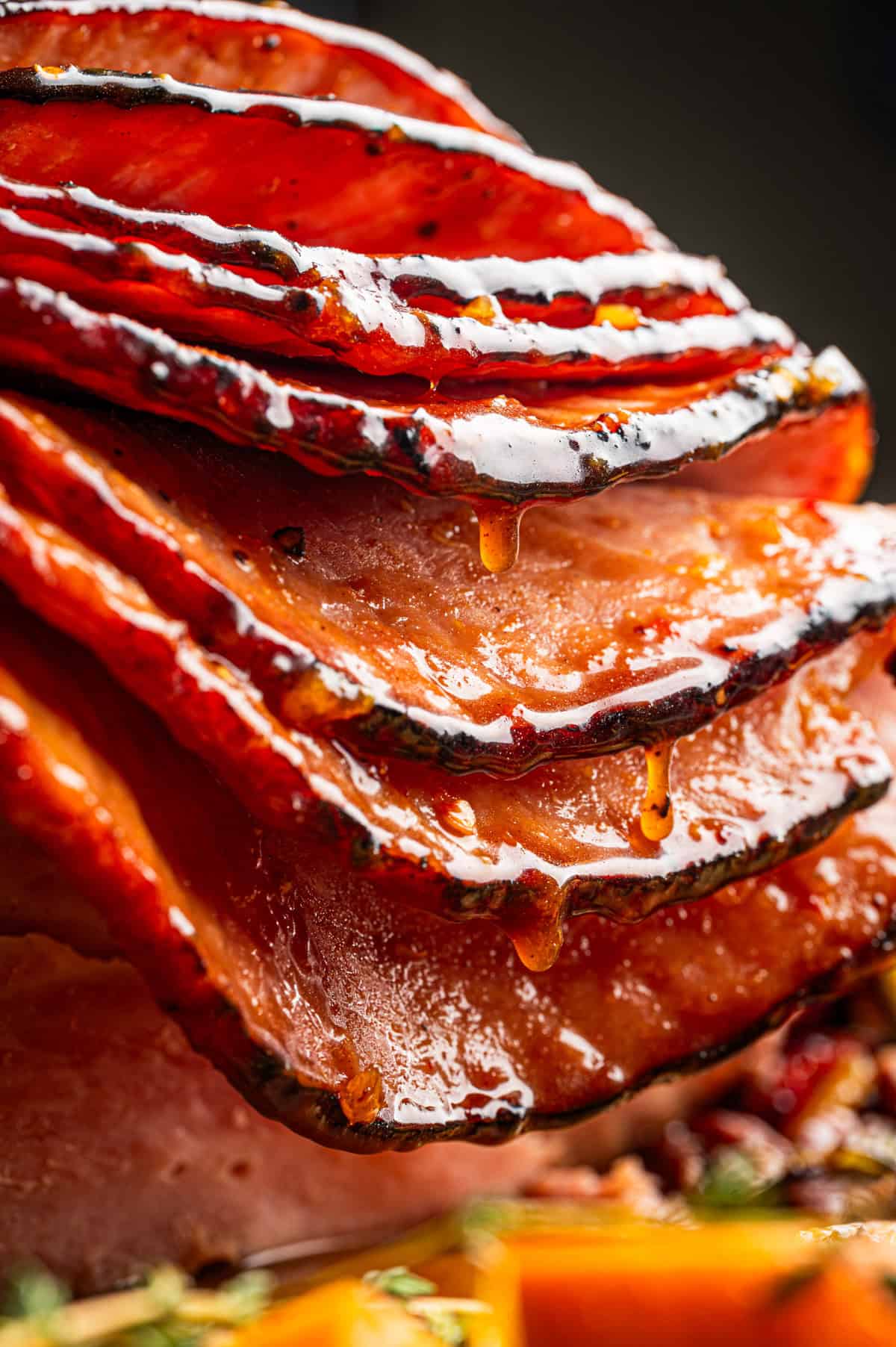 Close-up of several slices of glazed, baked and broiled ham stacked together, with caramelized edges and juices dripping down.