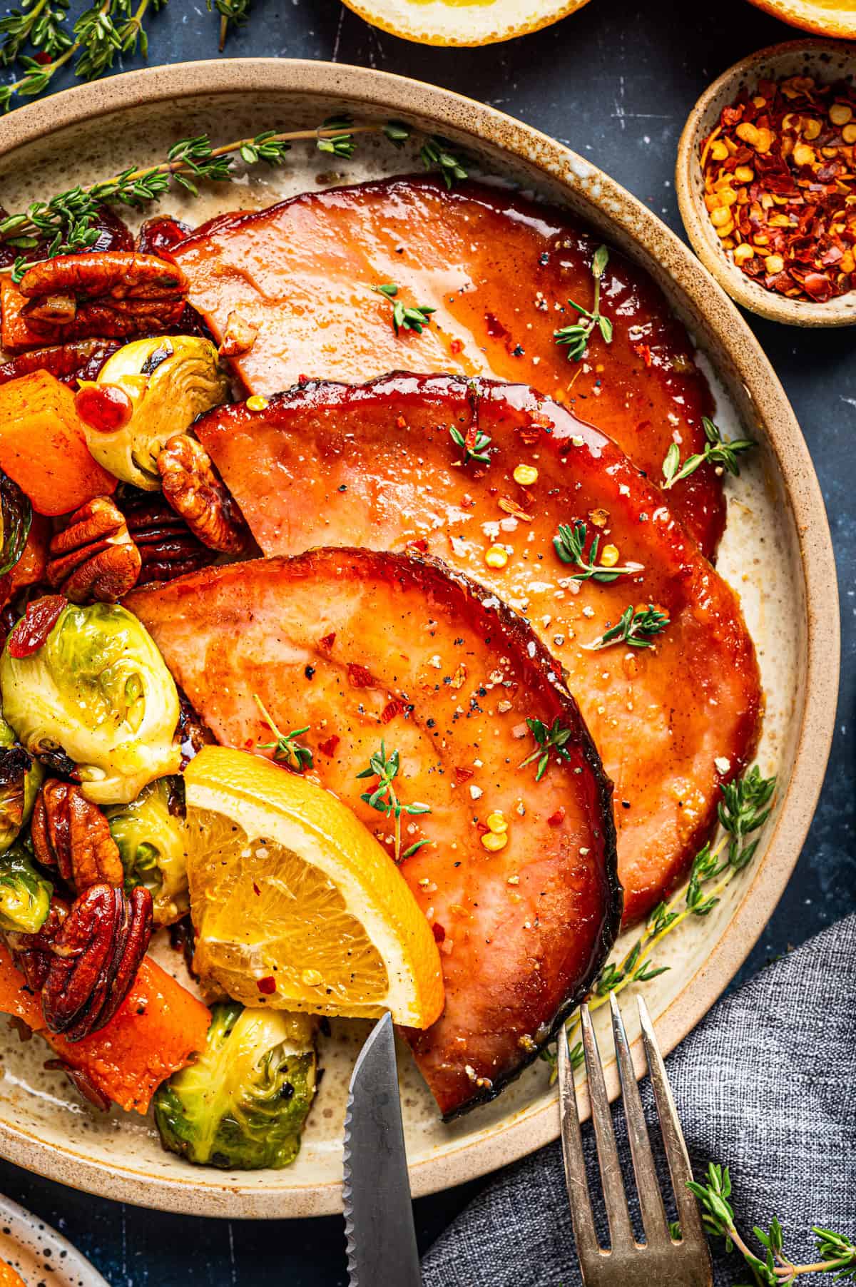 A plate with three glazed ham slices topped with herbs, a wedge of orange, roasted Brussels sprouts, pecans, and orange chunks. 