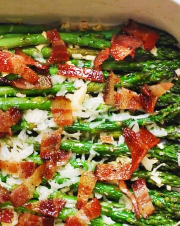 Fresh asparagus spears in a baking dish topped with crispy bacon pieces and melted shredded cheese.