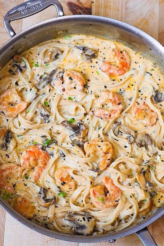 A skillet filled with creamy pasta topped with shrimp, sliced mushrooms, and herbs, all coated in a rich sauce with visible noodles and pieces of seafood.