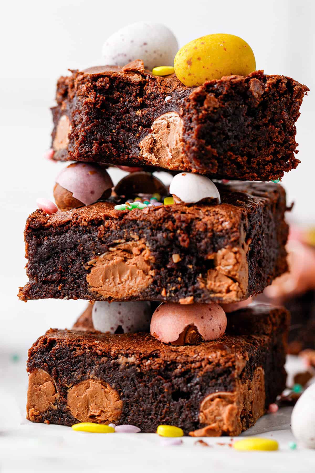 A stack of three rich chocolate brownies filled with chocolate chunks, decorated with colorful candy-coated chocolate eggs and sprinkles. The brownies appear moist and fudgy.