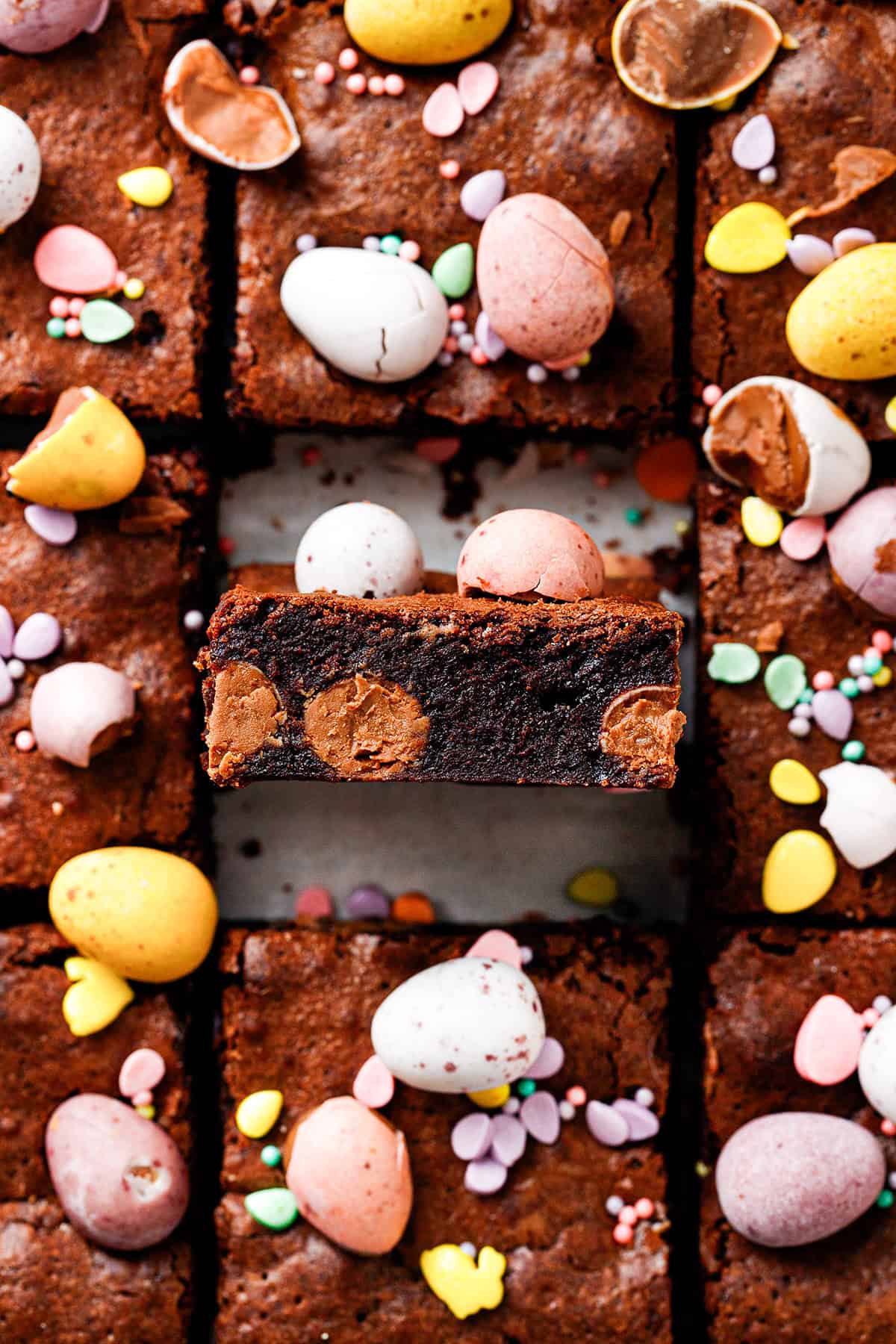 A close-up of chocolate brownies topped with colorful candy eggs and pastel sprinkles. One brownie is lifted to show its fudgy texture and chocolate chips inside, surrounded by more decorated brownies.