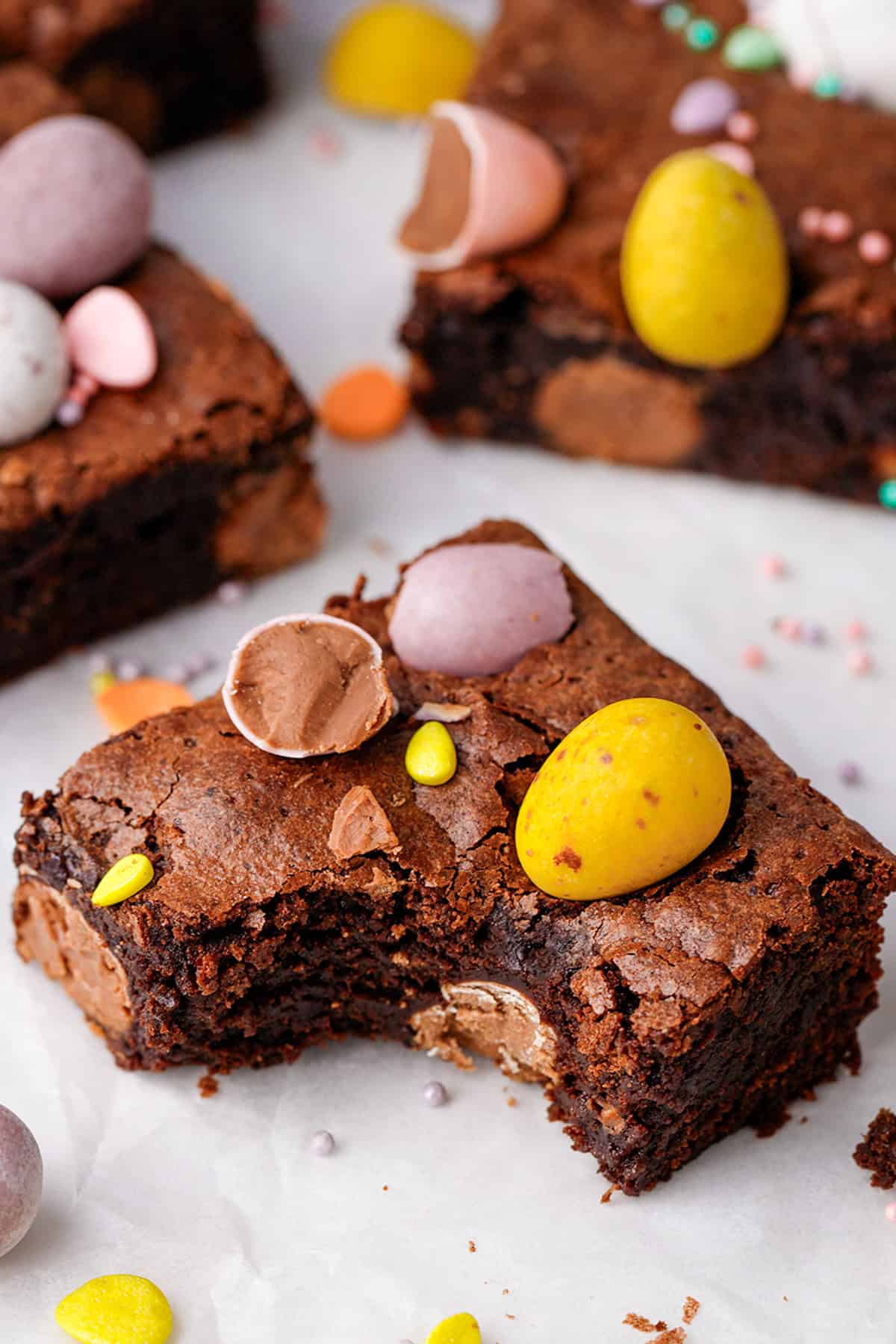 A close-up of chocolate brownies topped with colorful candy-coated chocolate eggs. One brownie has a bite taken out of it, revealing the rich, fudgy interior. Scattered candy pieces and sprinkles decorate the surface.