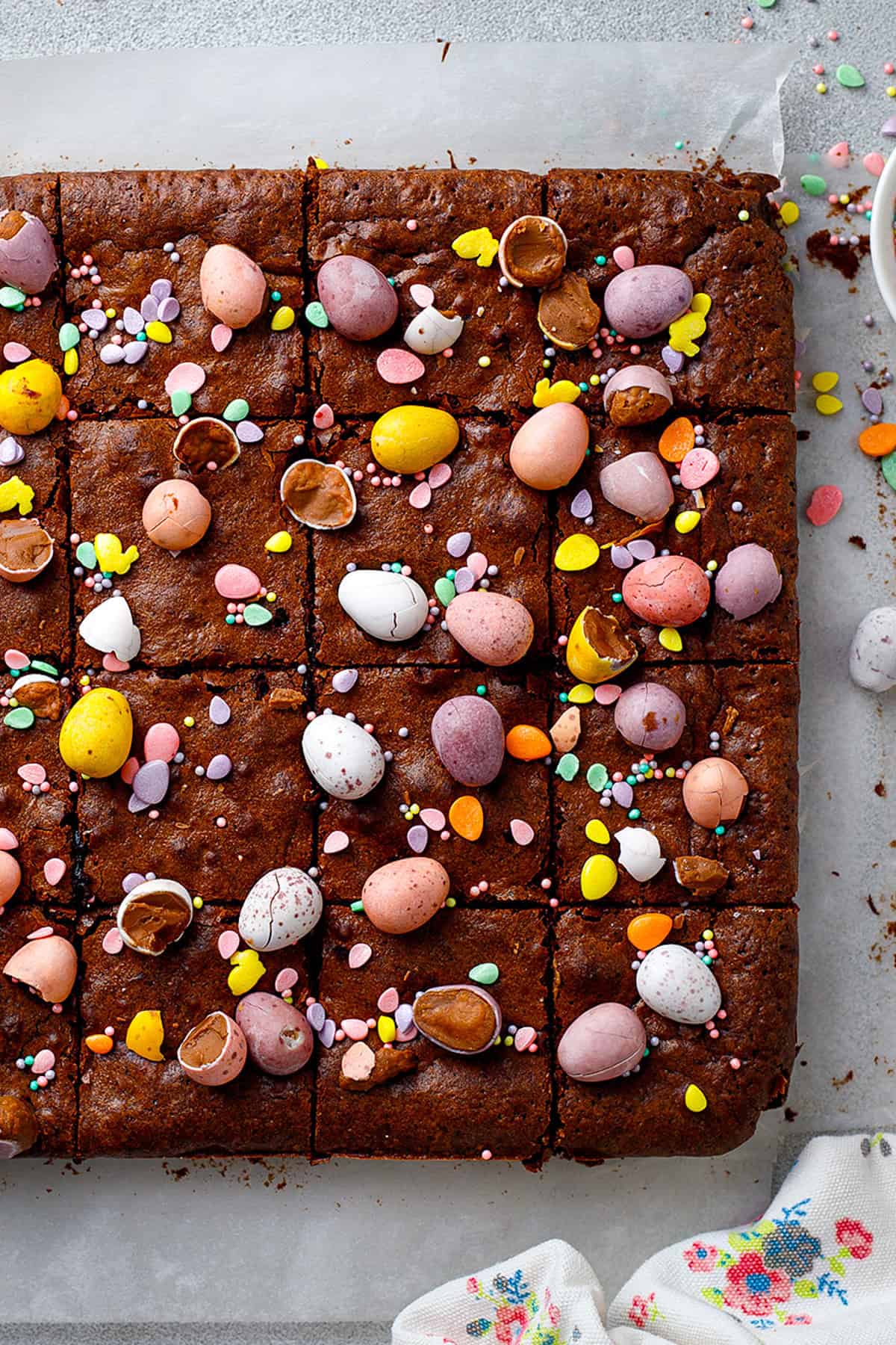 A tray of chocolate brownies cut into squares, topped with colorful candy-coated chocolate eggs, crushed egg pieces, and sprinkles, on parchment paper with a floral napkin nearby.