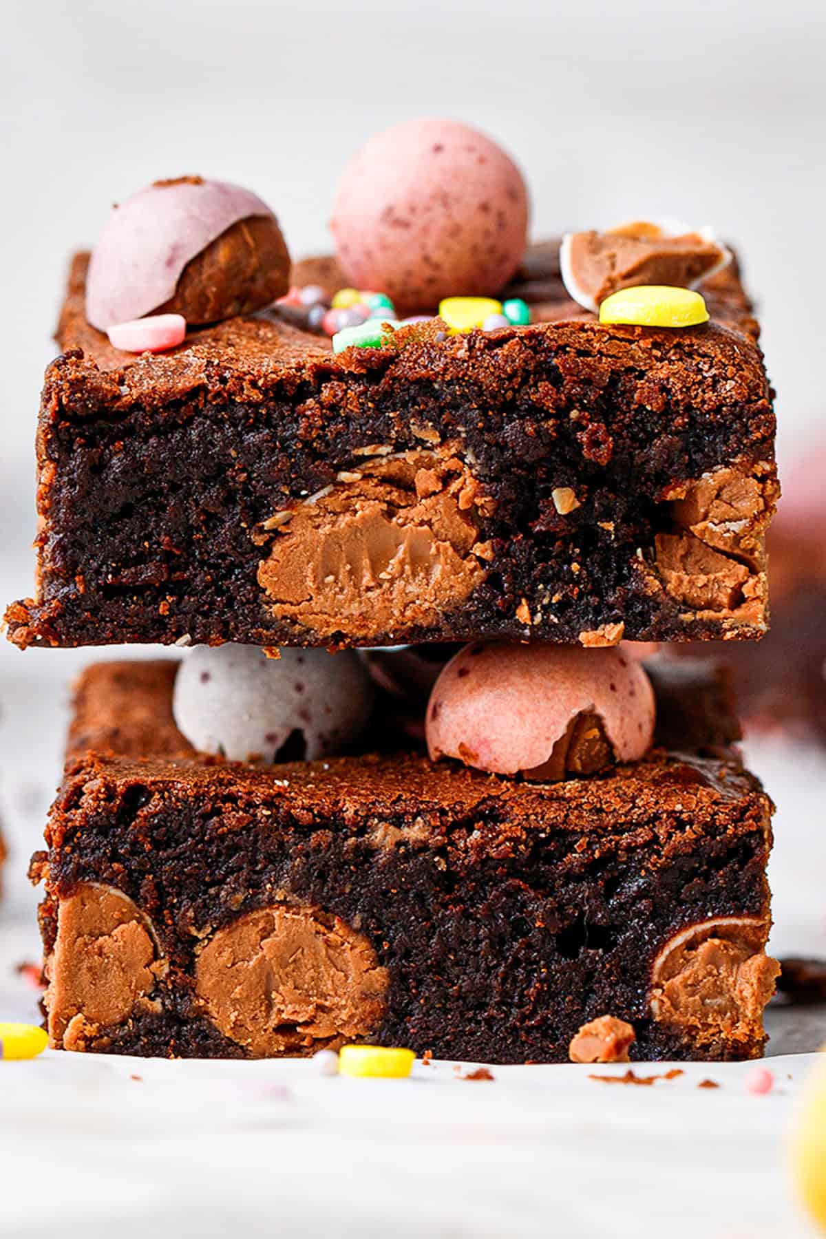 Two thick, fudgy chocolate brownies with visible chunks of chocolate are stacked. The top brownie is decorated with pastel candy eggs and colorful sprinkles. 