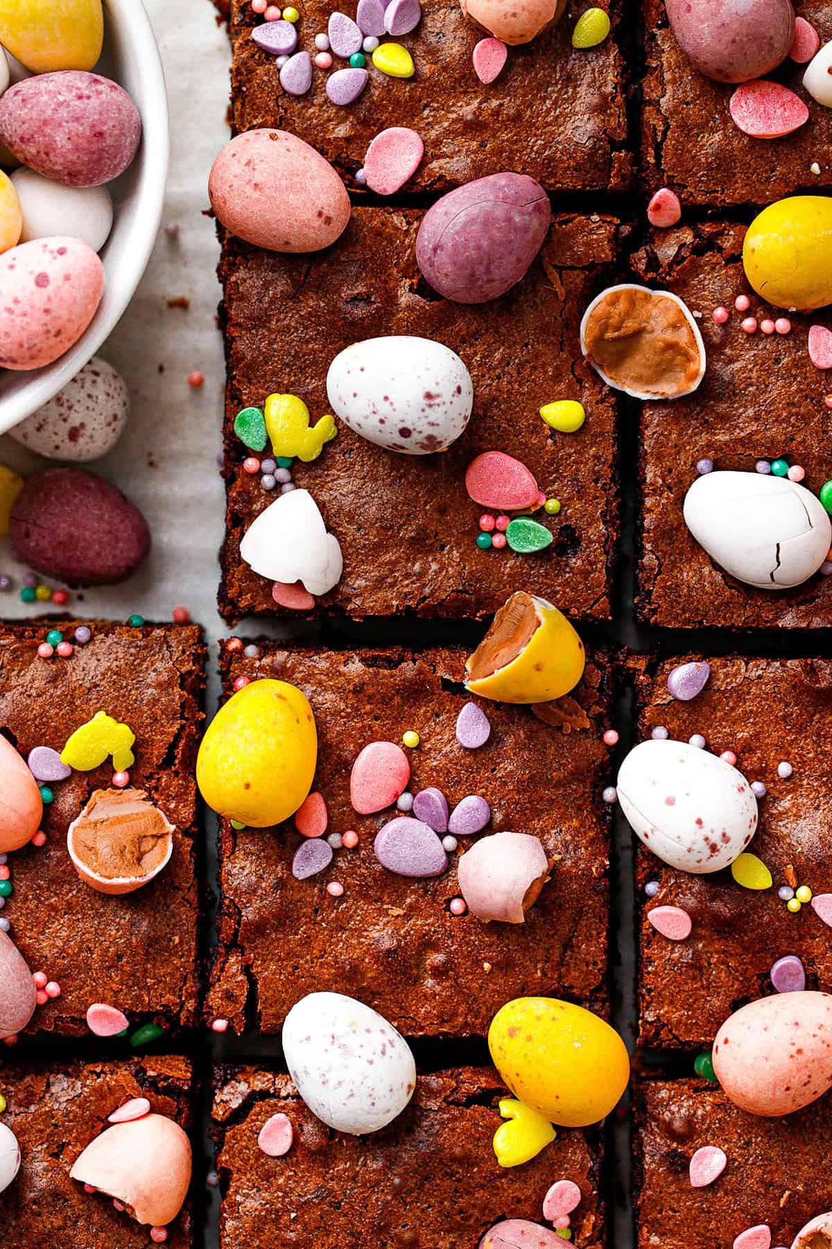 Brownies cut into squares are topped with colorful candy eggs, some whole and some cracked open. A bowl of additional candy eggs is visible in the top left corner. Small heart-shaped sprinkles are scattered over the brownies.
