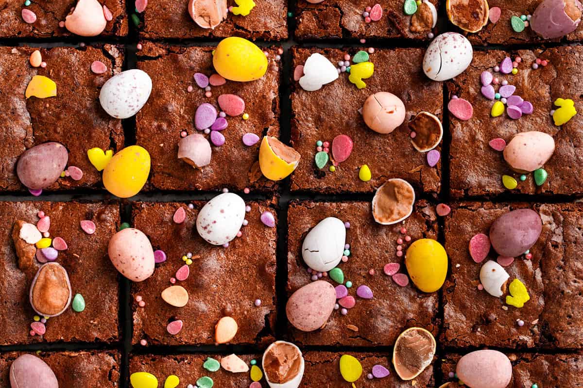 Brownie squares topped with colorful candy eggs, sprinkles, and broken eggshell pieces, arranged in a grid pattern to create a festive, playful look.