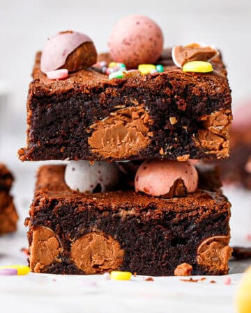 Two thick, fudgy chocolate brownies with visible chocolate chunks are stacked on top of each other. Both are topped with colorful candy-coated chocolate eggs and sprinkles, resting on a white surface with more candies scattered around.