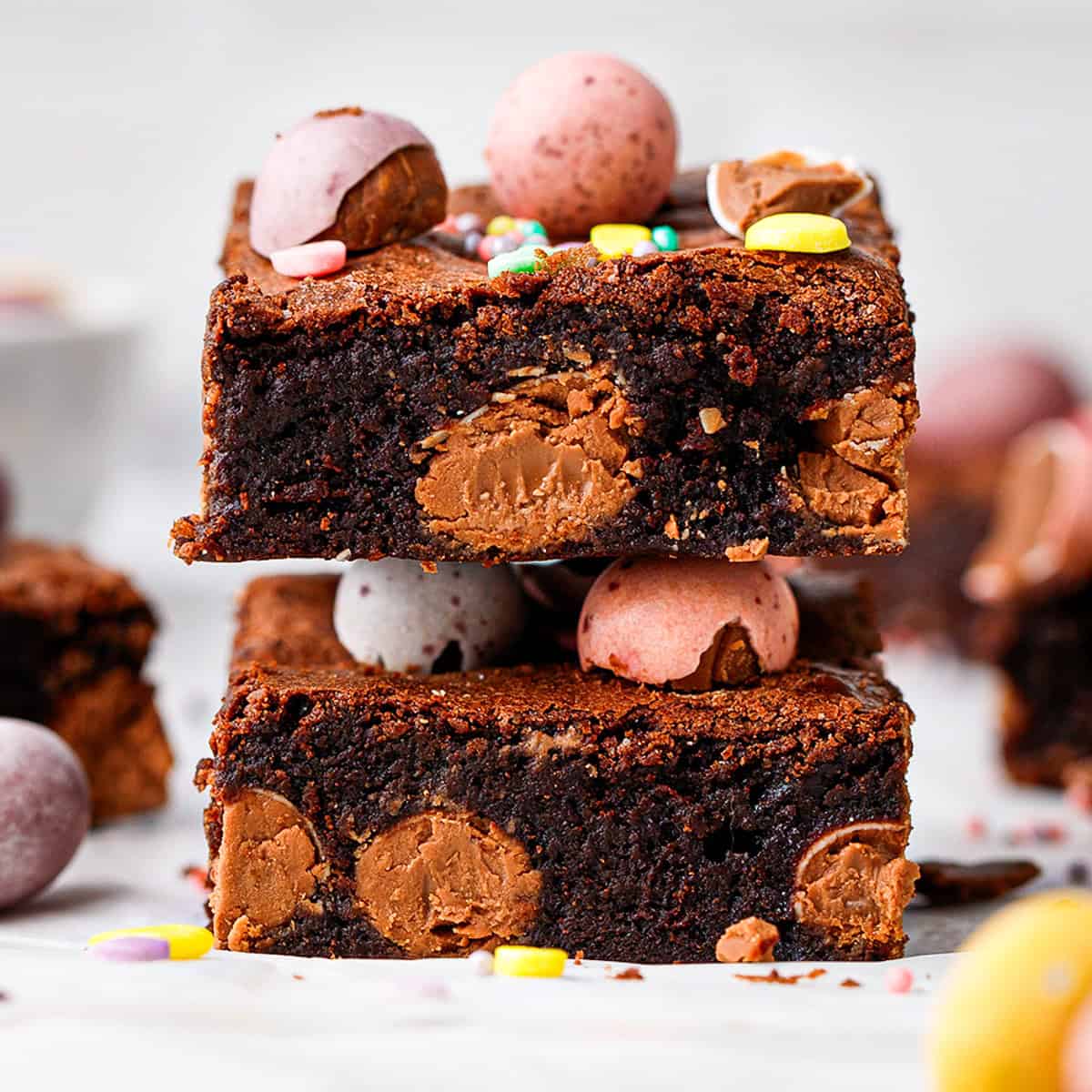 Two thick, fudgy chocolate brownies with visible chocolate chunks are stacked on top of each other. Both are topped with colorful candy-coated chocolate eggs and sprinkles, resting on a white surface with more candies scattered around.