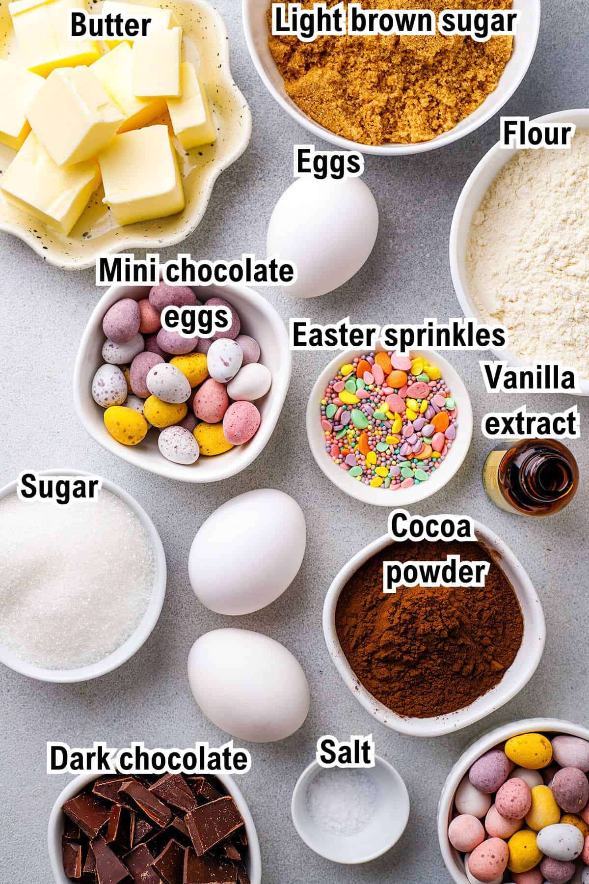 Various baking ingredients for Easter Brownies (labeled): butter, light brown sugar, flour, eggs, mini chocolate eggs, sugar, Easter sprinkles, vanilla extract, cocoa powder, dark chocolate, and salt, arranged in bowls on a countertop.
