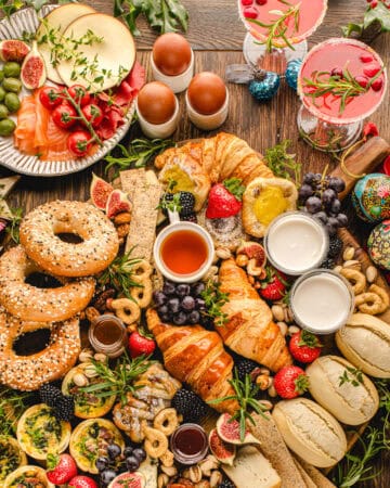 A festive brunch spread with bagels, croissants, bread rolls, smoked salmon, cheeses, figs, berries, soft-boiled eggs, quiche, dips, fruits, and pink cocktails, decorated with ornaments and holiday greenery.