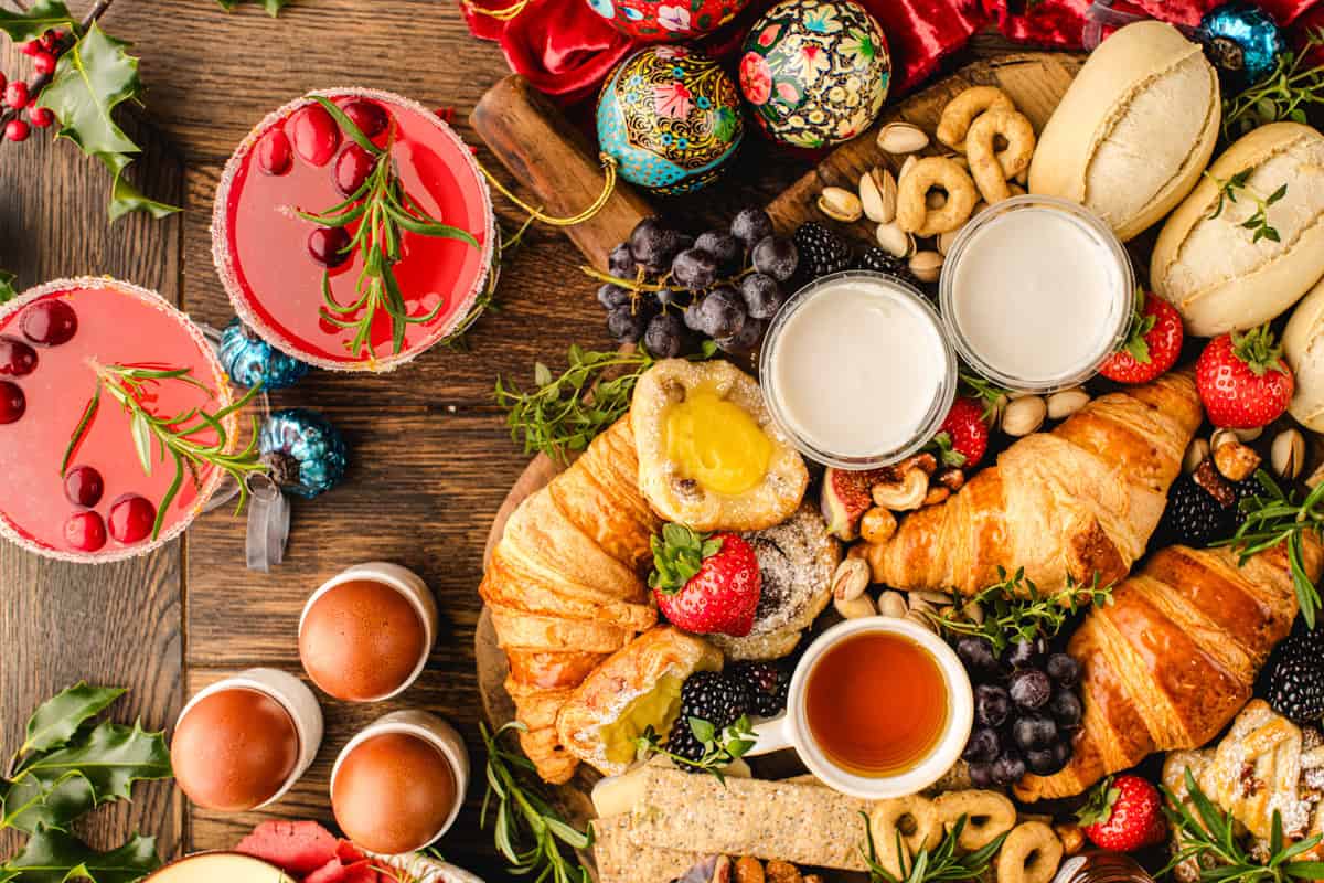 A festive breakfast spread with croissants, pastries, fruit, boiled eggs, tea, pink cranberry drinks on a wooden table.