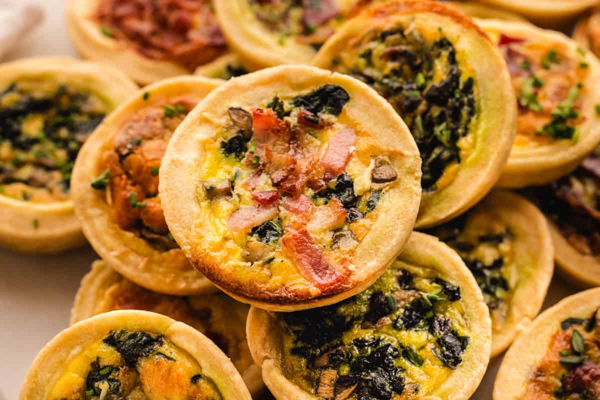 A close-up of several mini quiches with golden crusts, topped with ingredients like spinach, bacon, and mushrooms, piled together on a platter.