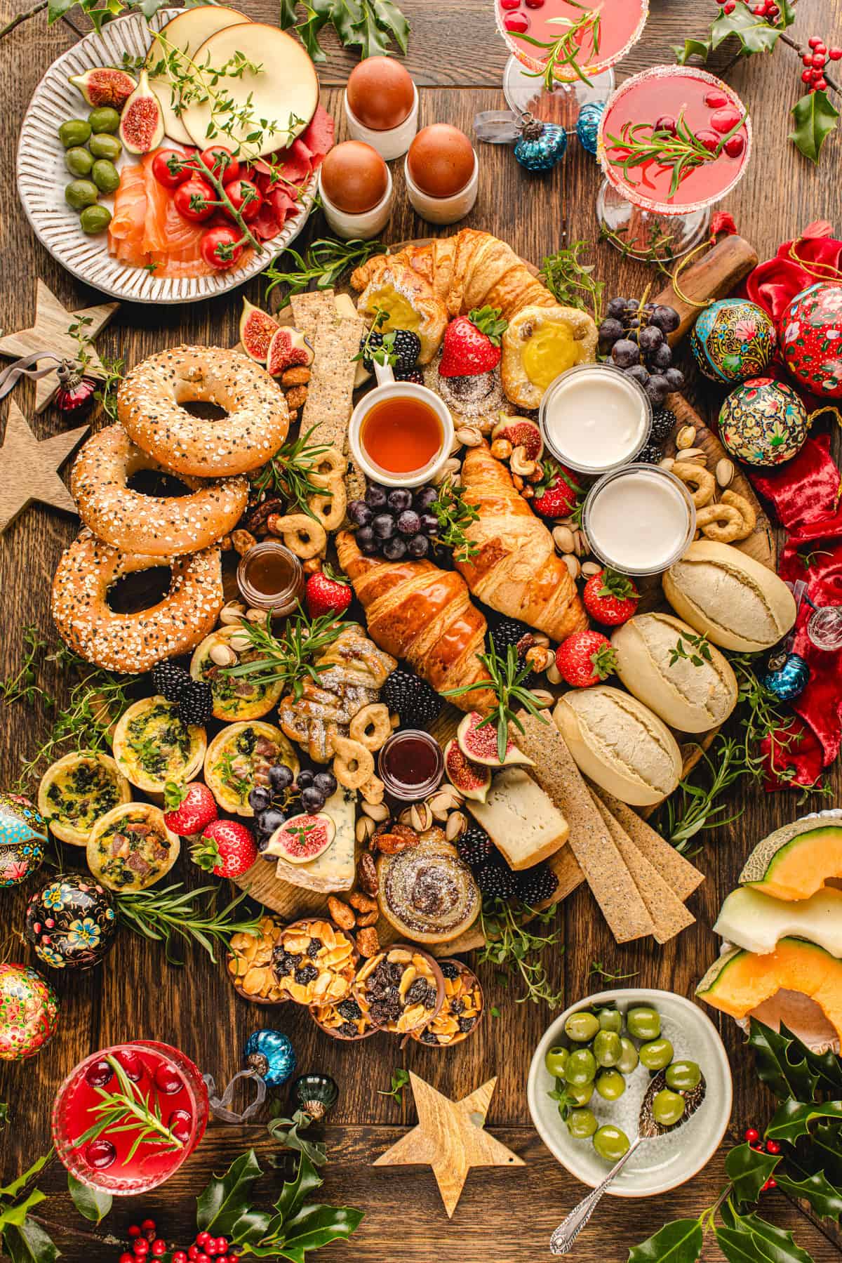 A festive breakfast spread on a wooden table features bagels, croissants, pastries, fruit, nuts, olives, jam, colorful eggs, drinks.