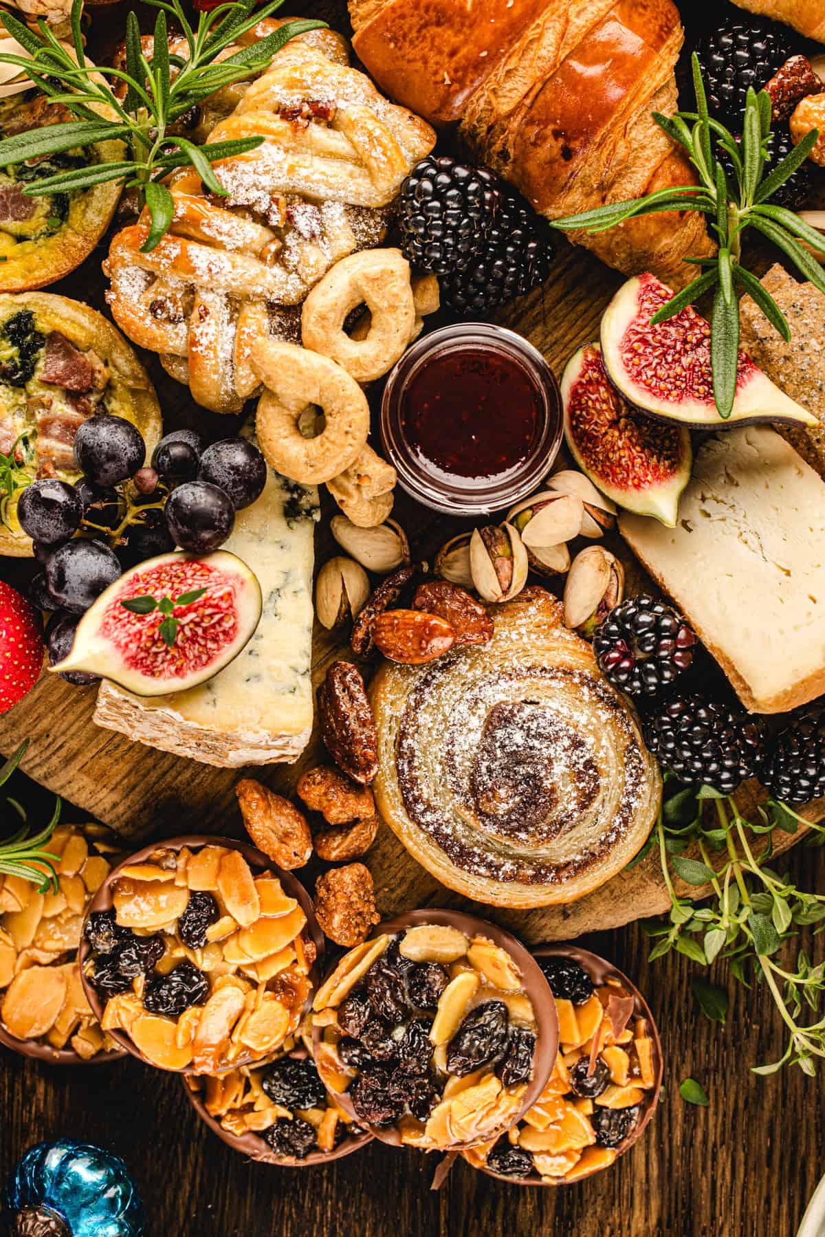 A wooden platter filled with assorted pastries, cheeses, figs, grapes, berries, cookies, nuts, and a small jar of jam, arranged decoratively with fresh herbs.