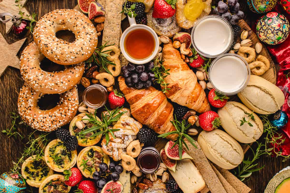 A colorful breakfast spread with bagels, croissants, bread rolls, fruit, pastries, cookies, jams, quiches, nuts, and cups of tea or coffee, arranged on a rustic wooden table with herbs and decorated eggs.