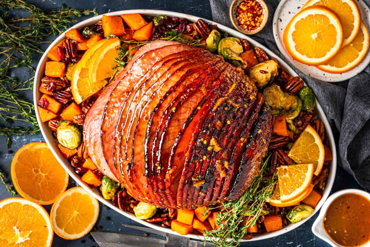 20 Best Easter Dinner Ideas (Main Dishes) A glazed spiral ham on a platter, surrounded by roasted Brussels sprouts, sweet potatoes, pecans, orange slices, and fresh herbs. Small bowls of orange slices and crushed red pepper are next to the platter.