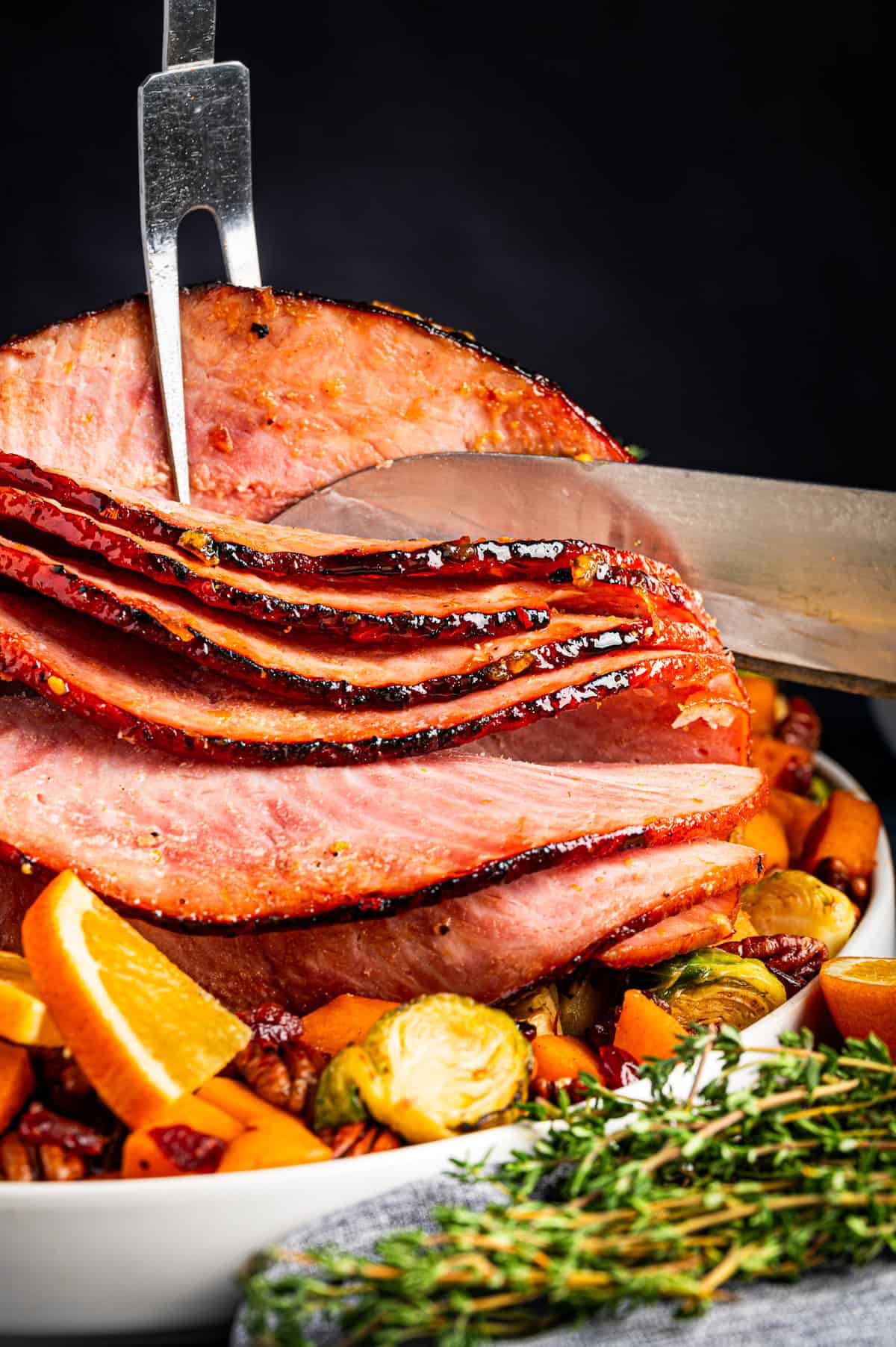Sliced glazed ham being carved with a knife and fork, served on a platter with roasted Brussels sprouts, orange slices, and herbs.