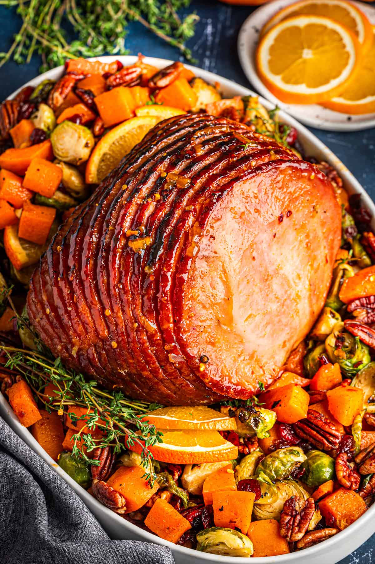A glazed baked ham garnished with orange slices, fresh thyme, and surrounded by roasted vegetables, including Brussels sprouts, pecans, and chunks of butternut squash on a serving platter.