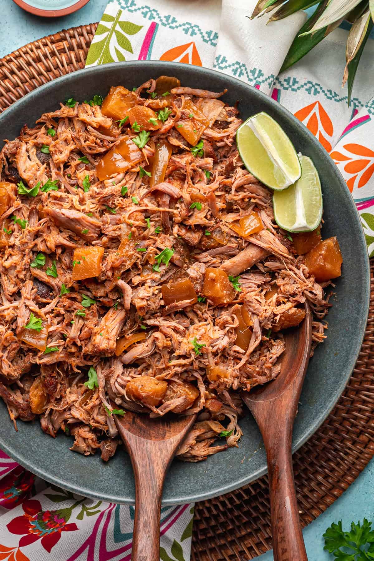 A large bowl filled with kalua pork and chunks of pineapple, garnished with chopped herbs and lime wedges, with two wooden serving spoons on a colorful patterned tablecloth.