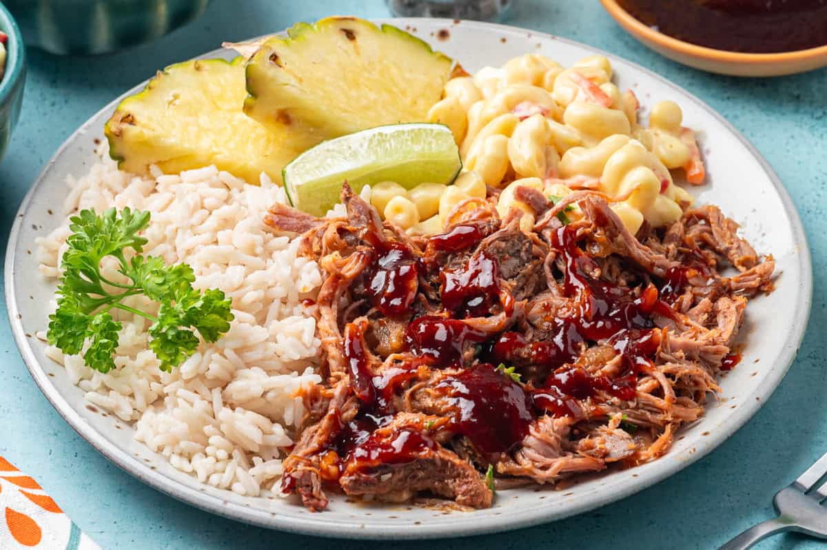 A plate of white rice, tender kalua pork with barbeque sauce, macaroni salad, pineapple slices, and a lime wedge, garnished with a sprig of parsley.