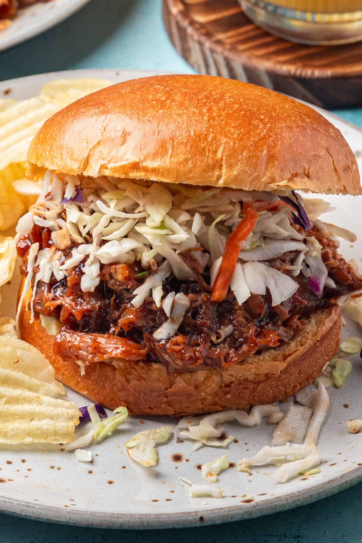 A kalua pork sandwich topped with coleslaw on a brioche bun, served on a plate with potato chips.