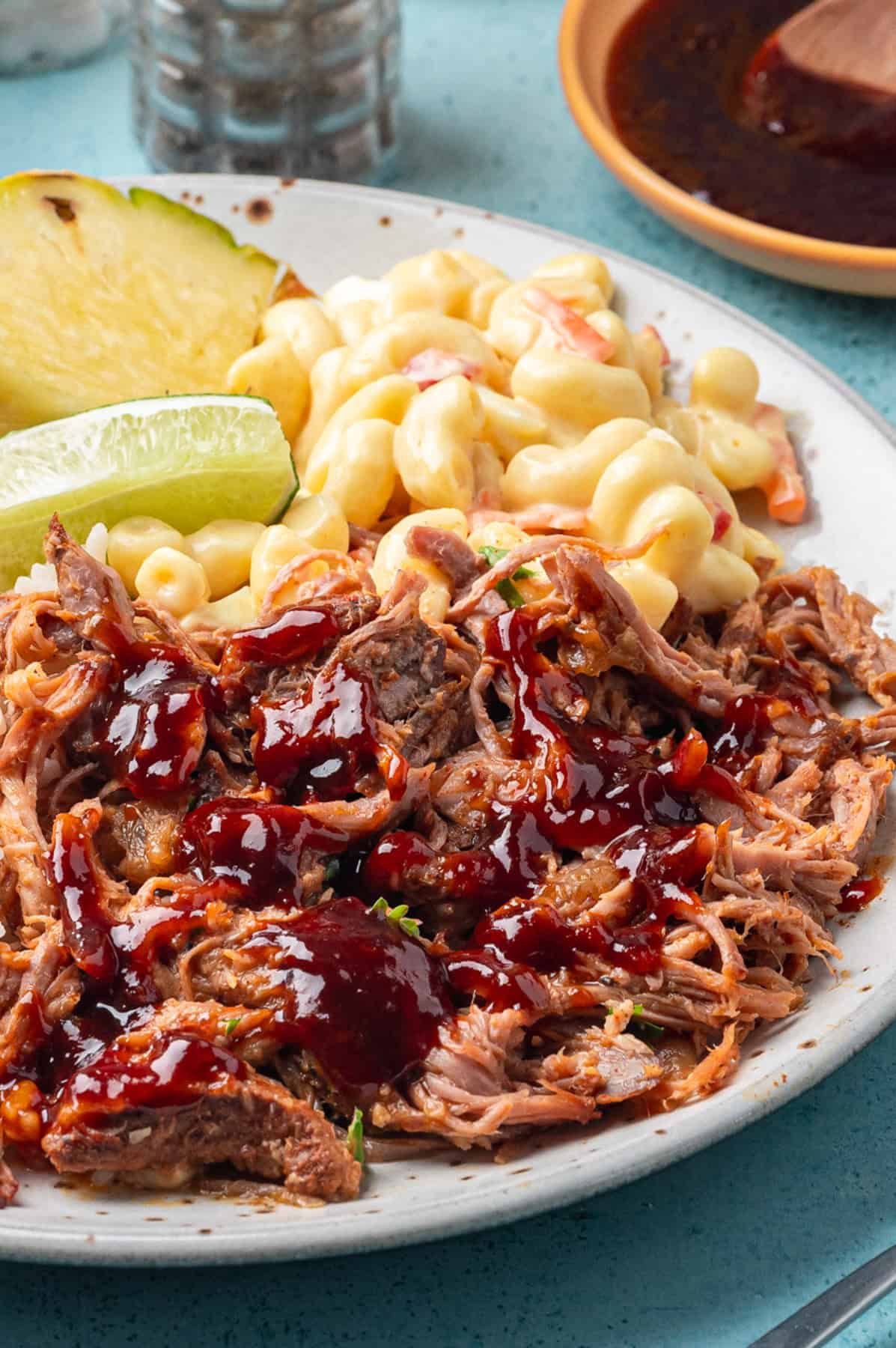 A plate with shredded kalua pork topped with sauce, creamy macaroni salad, and slices of lime and pineapple, set on a blue tabletop.