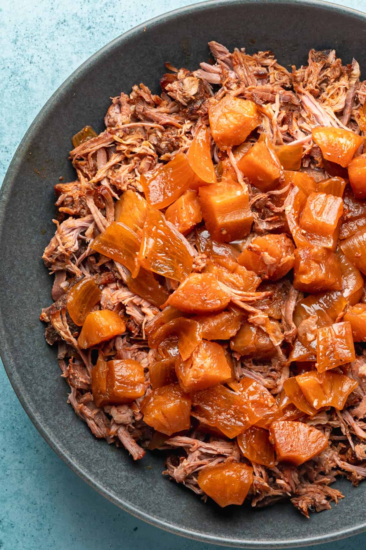 Kalua pork topped with cooked pineapple chunks and sauce, served in a dark bowl on a light blue surface.