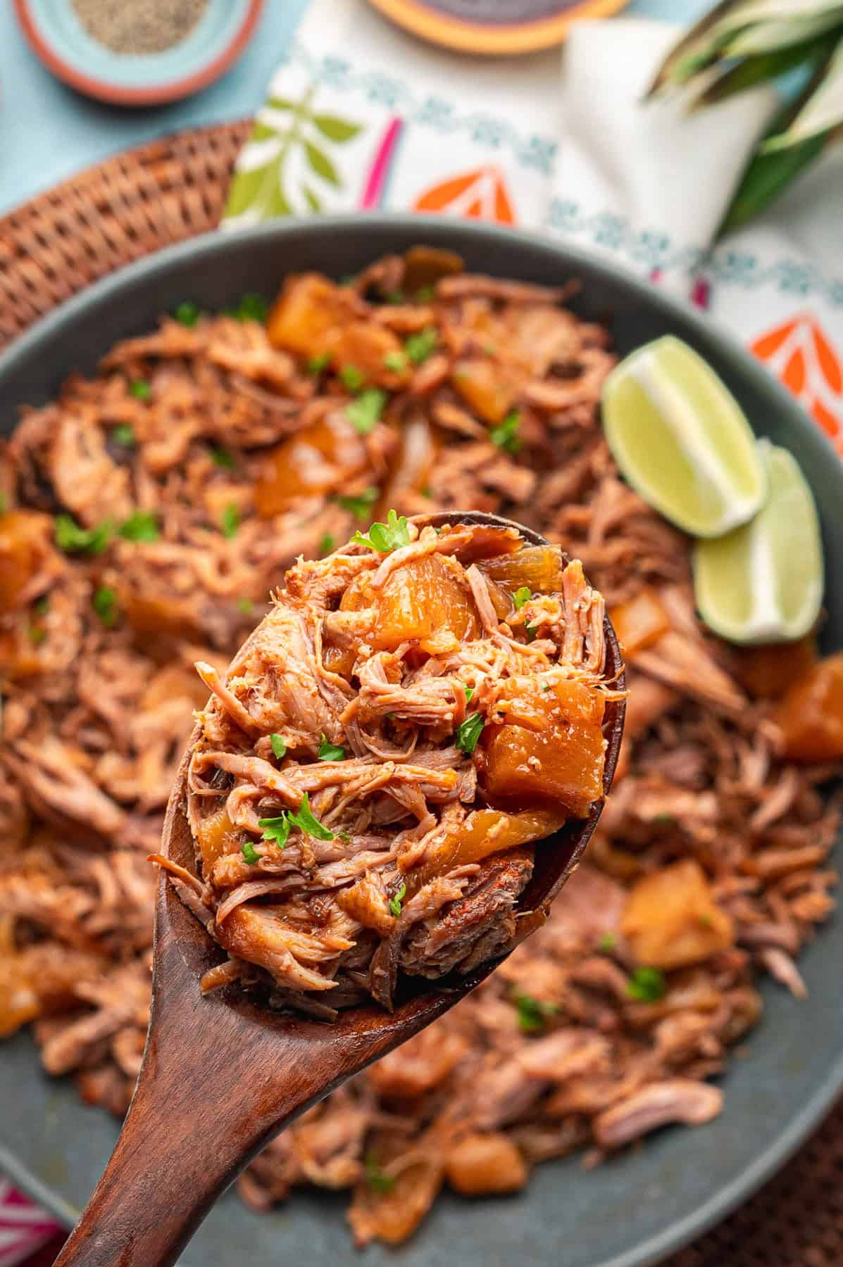 A wooden spoon holds a serving of kalua pork with chunks of pineapple, garnished with herbs. In the background, a large plate of the same dish and lime wedges are visible.