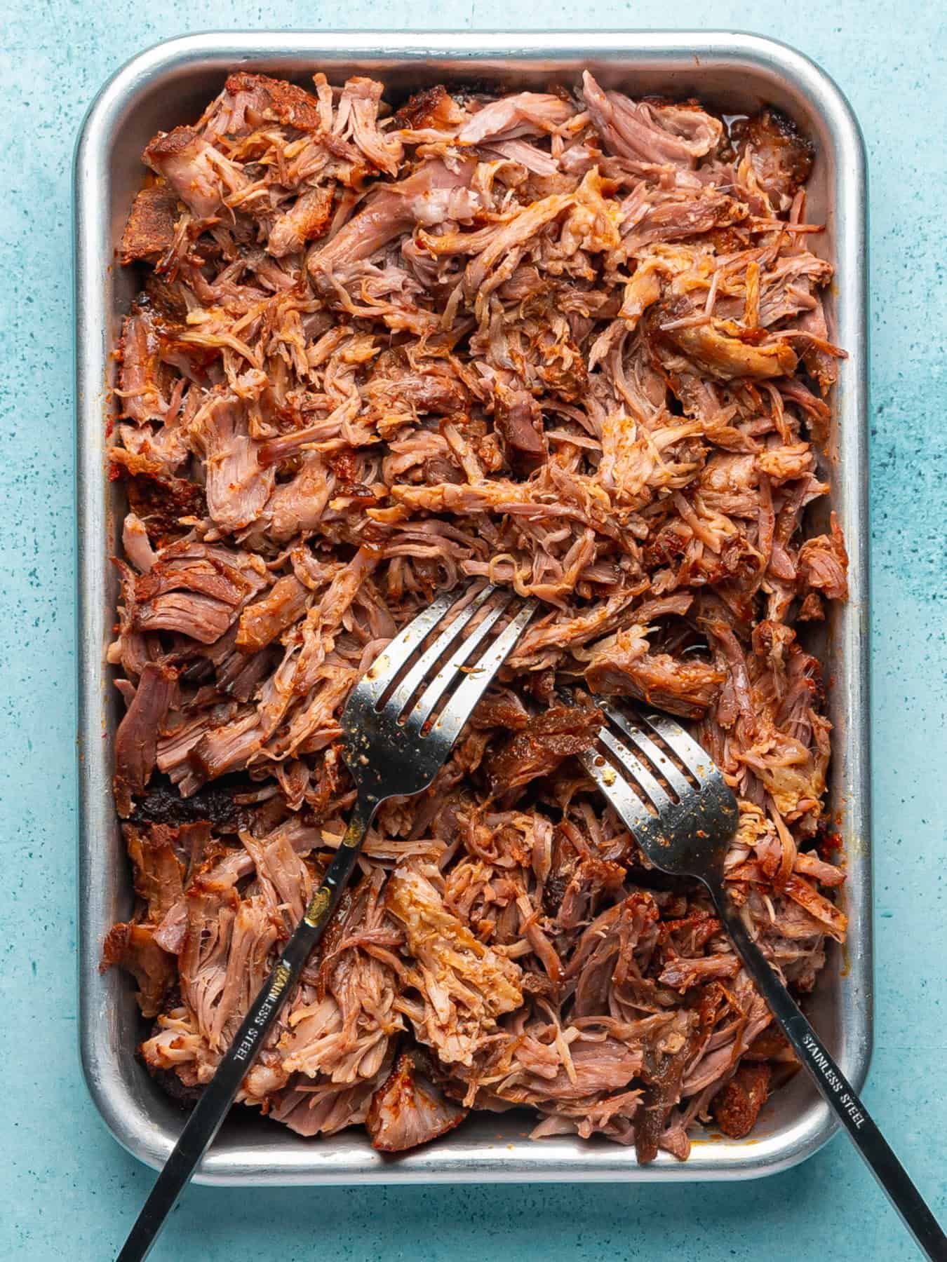 A baking tray filled with shredded, cooked kalua pork, with two metal forks resting on top for serving. The meat appears tender and juicy, with crispy edges. The background is a light blue surface.