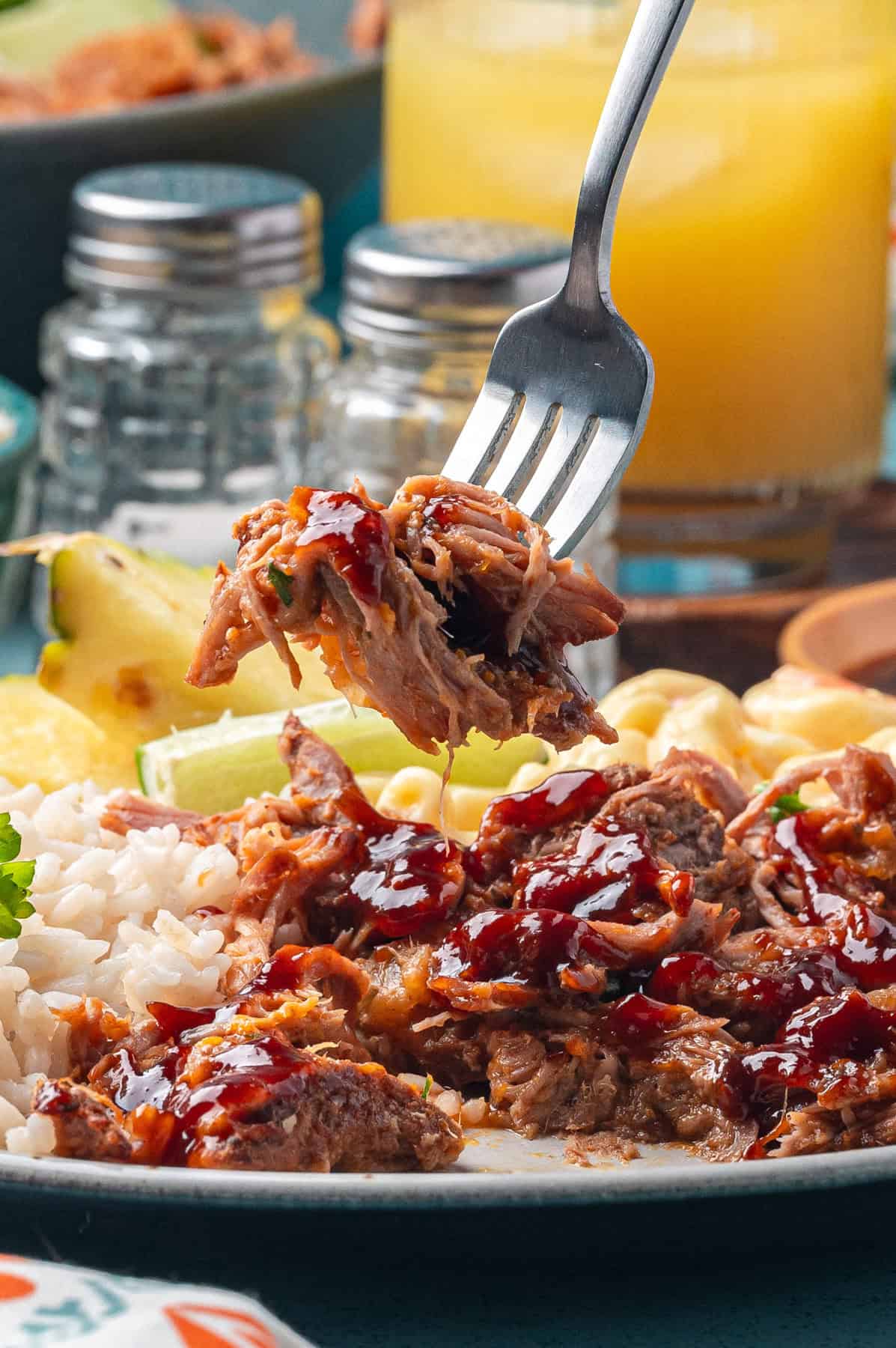 A close-up of a fork lifting tender kalua pork drizzled with sauce, served with white rice, pineapple, and corn on a plate. A glass of orange juice and salt and pepper shakers are in the background.