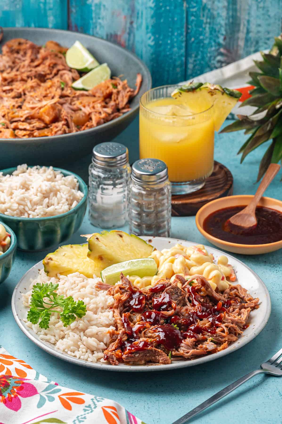 A plate with kalua pork topped with sauce, white rice, grilled pineapple, lime wedge, and macaroni salad. A bowl of rice, glass of pineapple juice, salt and pepper shakers, and a large bowl of shredded kalua pork are in the background.