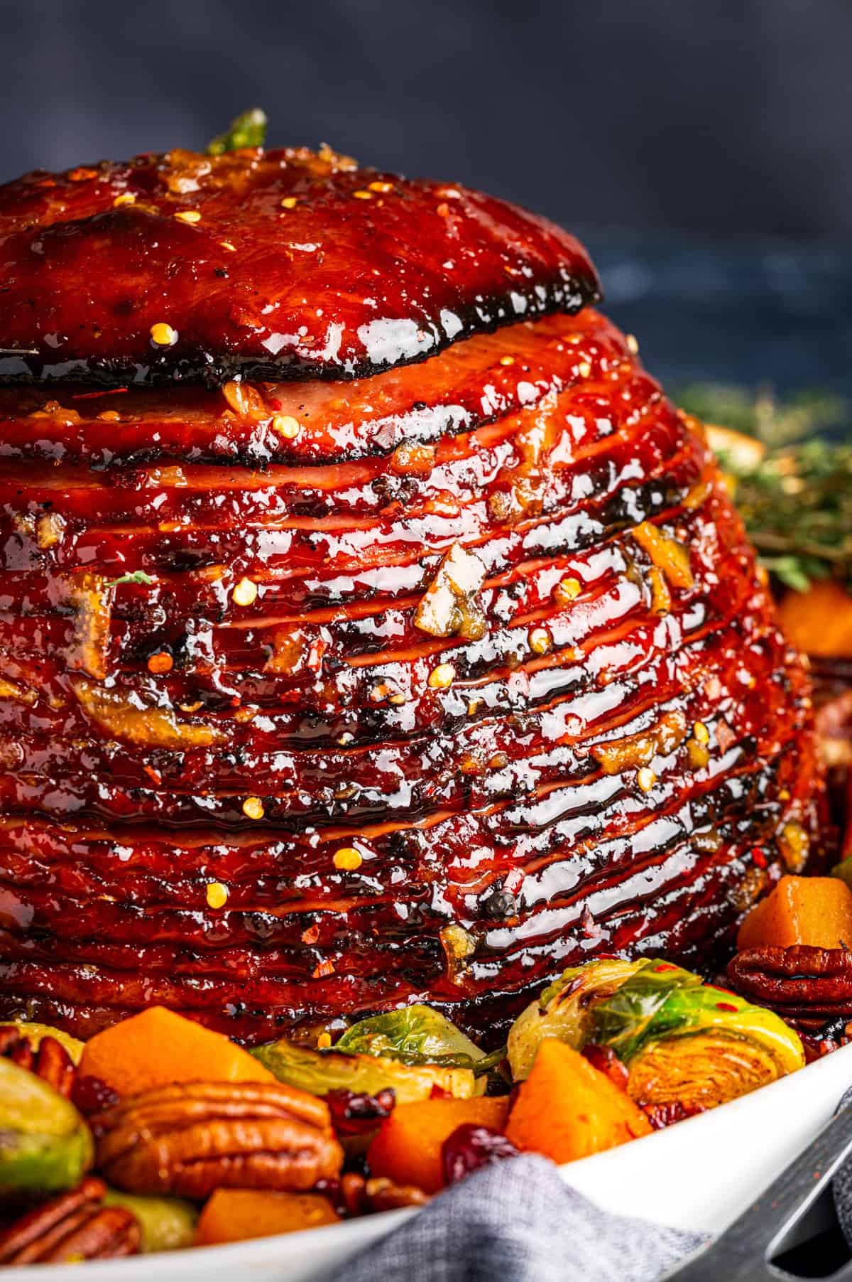 A close-up of a glazed spiral-cut ham, topped with herbs and surrounded by roasted vegetables, pecans, and cranberries on a white platter. The ham is shiny and caramelized with visible spices.