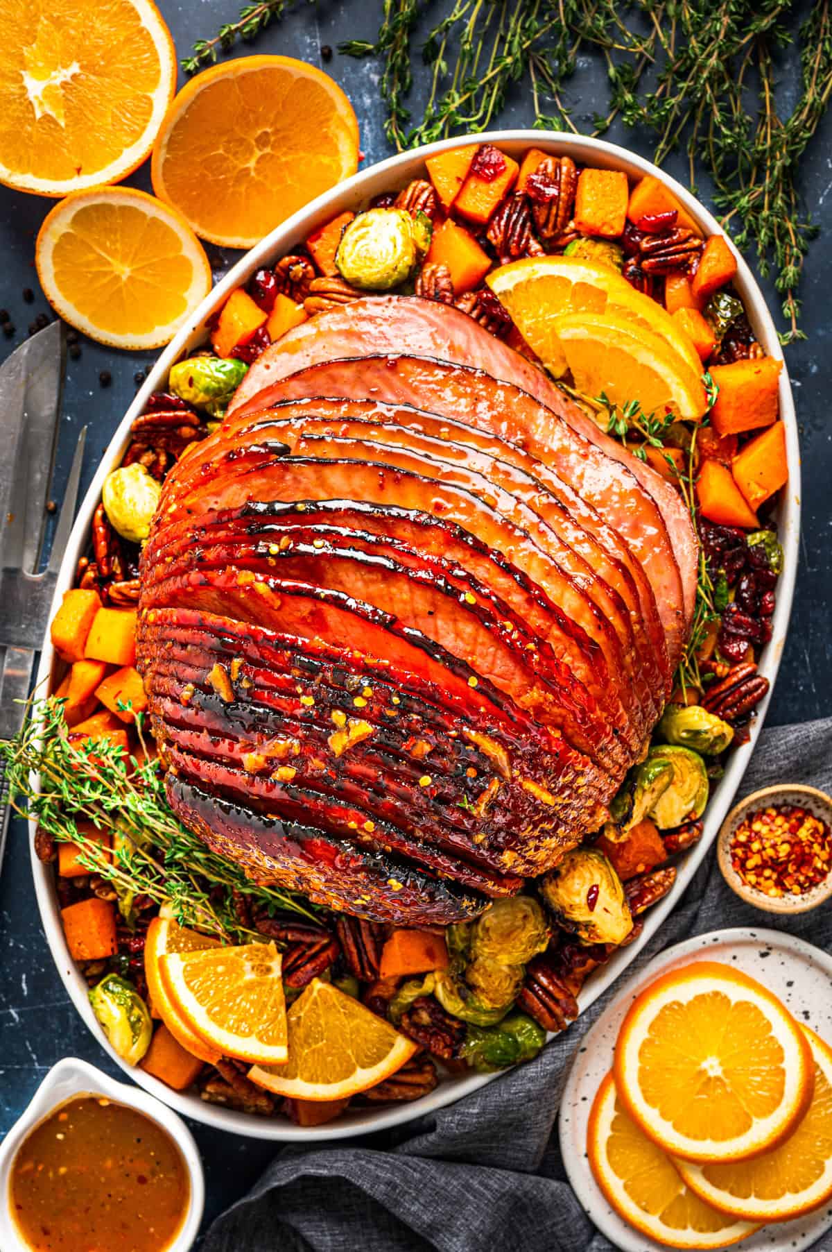 A glazed spiral ham garnished with fresh orange slices, pecans, Brussels sprouts, Butternut Squash, and herbs is served on an oval platter. Sliced oranges and a bowl of sauce sit nearby on a dark table.