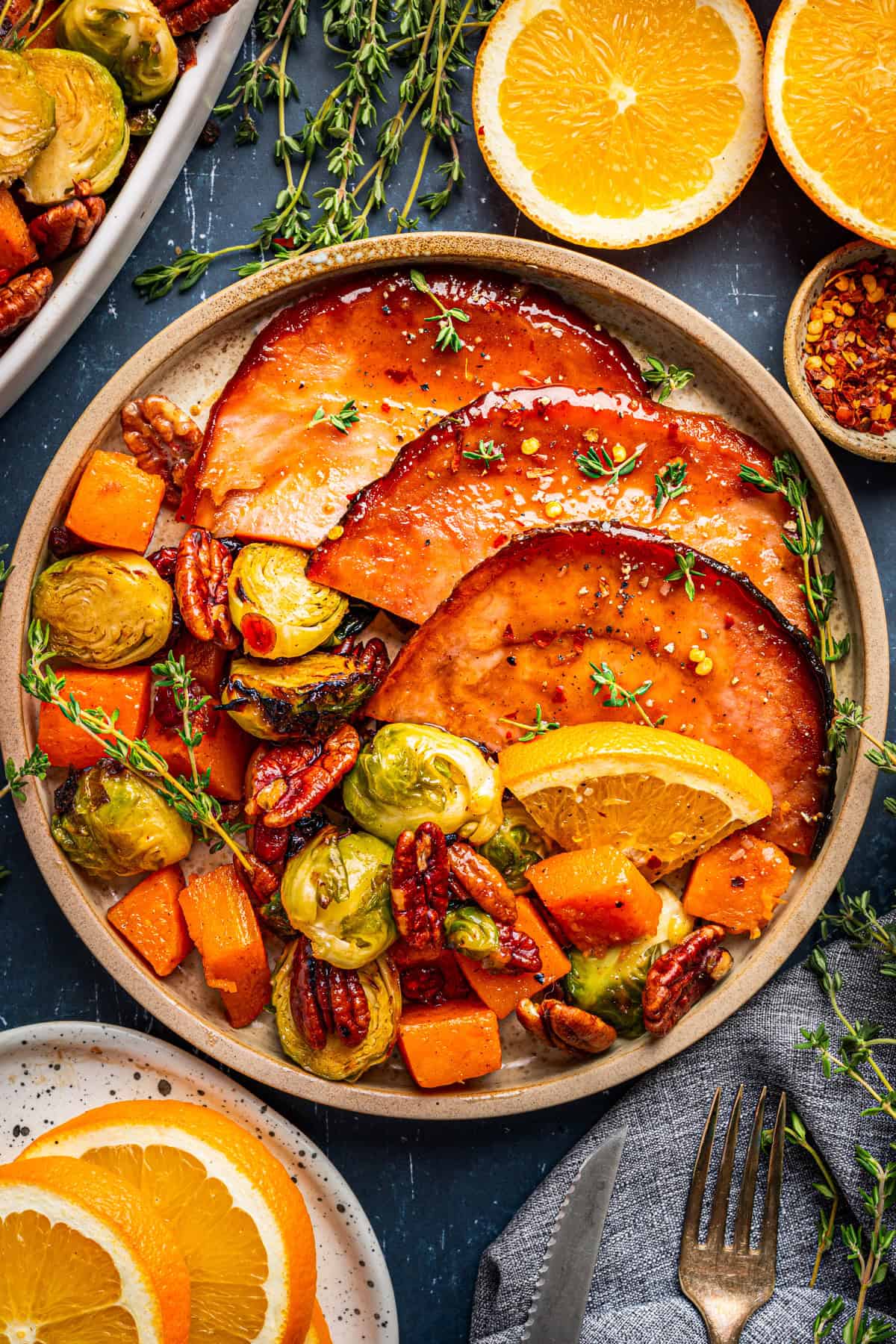 A plate with glazed ham slices, roasted Brussels sprouts, pecans, butternut squash, and orange wedges, garnished with fresh thyme. Halved oranges and extra serving plates are visible in the background.