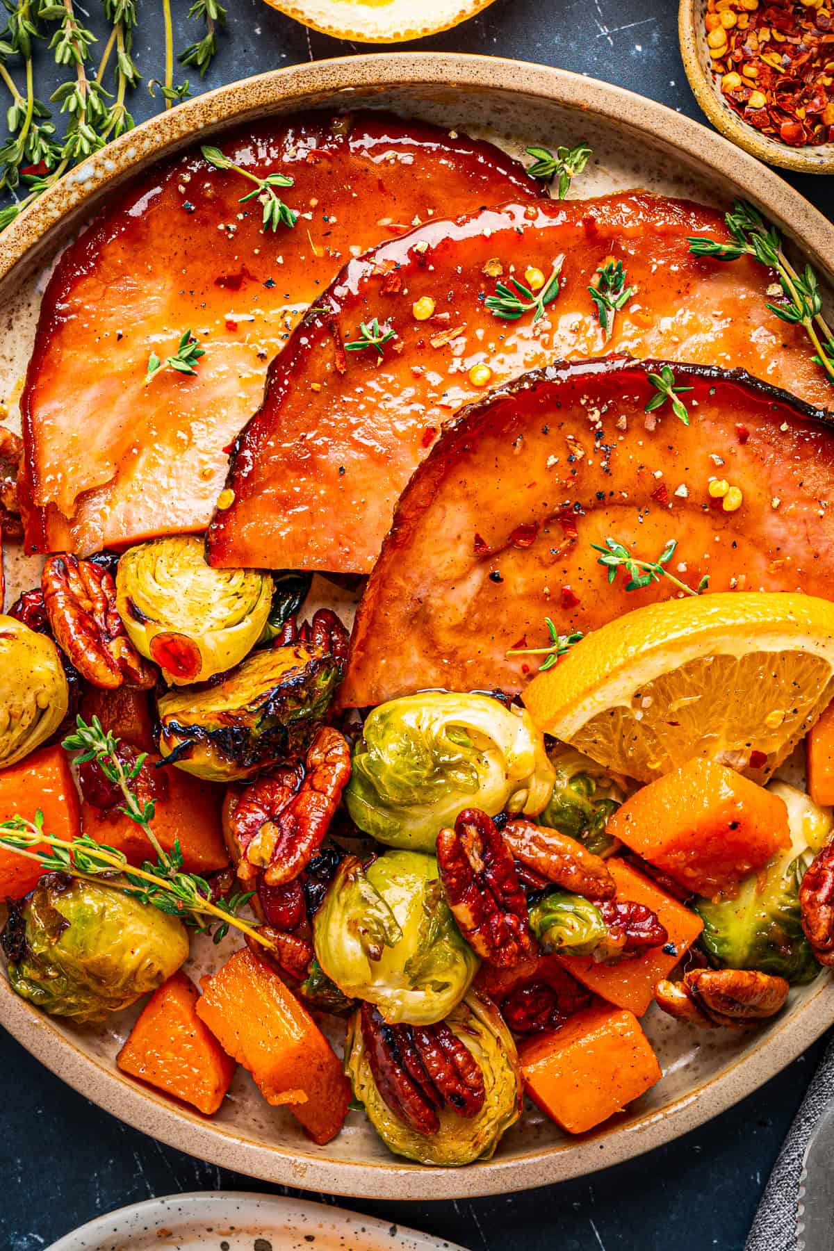 A plate with glazed ham slices, roasted Brussels sprouts, pecans, and butternut squash, garnished with fresh thyme and a lemon wedge.