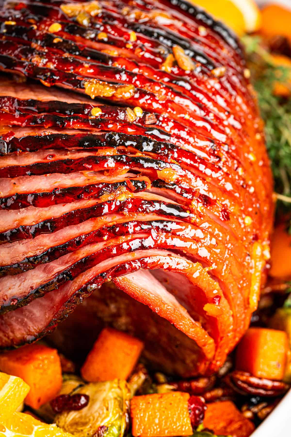 Close-up of a glazed, spiral-sliced ham with a shiny, caramelized surface, surrounded by roasted vegetables and pecans.