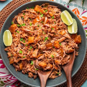 A bowl filled with shredded, seasoned kalua pork mixed with chunks of pineapple, garnished with chopped herbs and lime wedges, with two wooden serving spoons beside the dish.