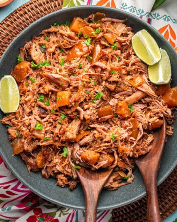 A bowl of kalua pork mixed with pineapple chunks, garnished with chopped herbs and lime wedges, served with two wooden serving spoons on a colorful patterned tablecloth.