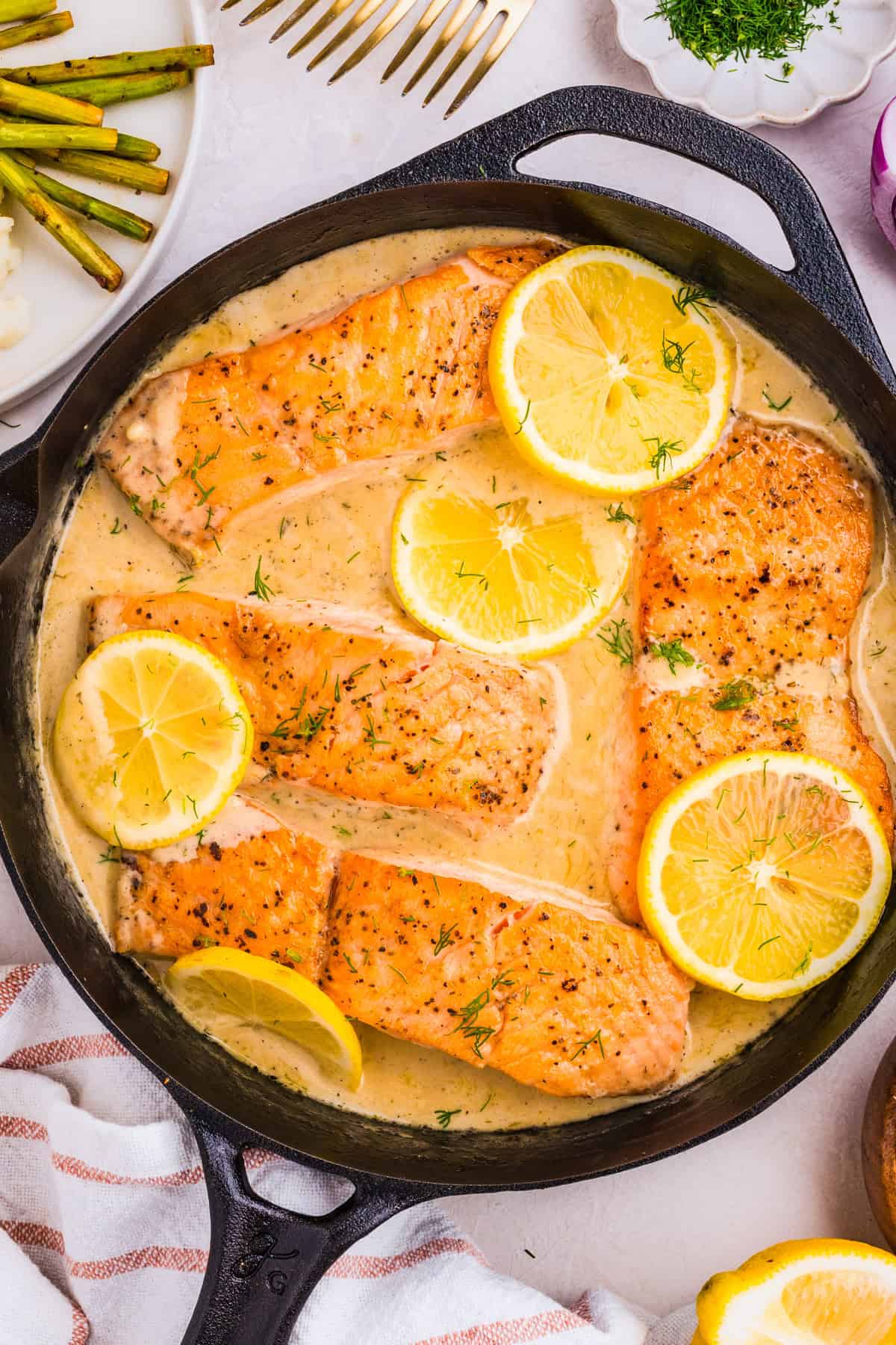 Four lemon dill salmon fillets cook in a creamy sauce in a cast iron skillet, garnished with fresh dill and lemon slices. 