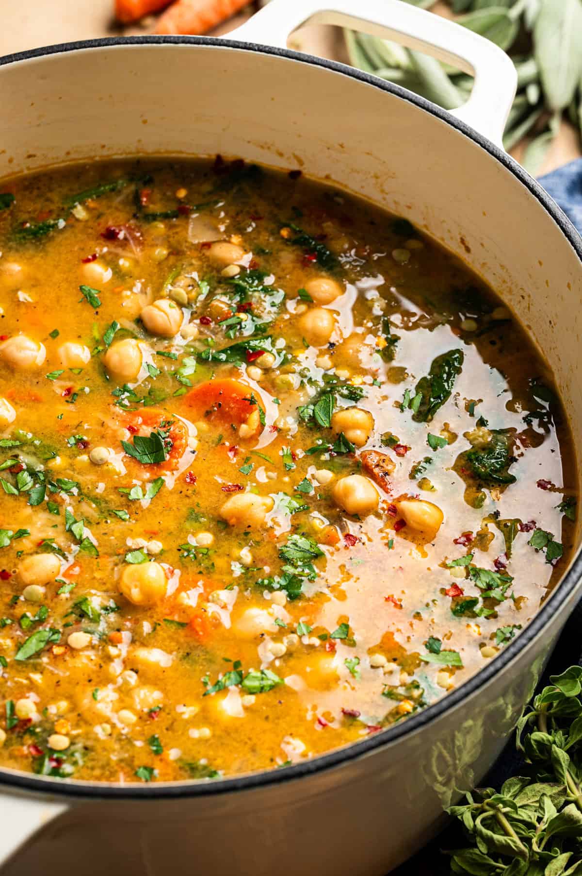 A close-up of a pot filled with vegetable lentil soup containing chickpeas, carrots, greens, herbs, and spices in a golden broth. Fresh herbs and vegetables surround the pot.