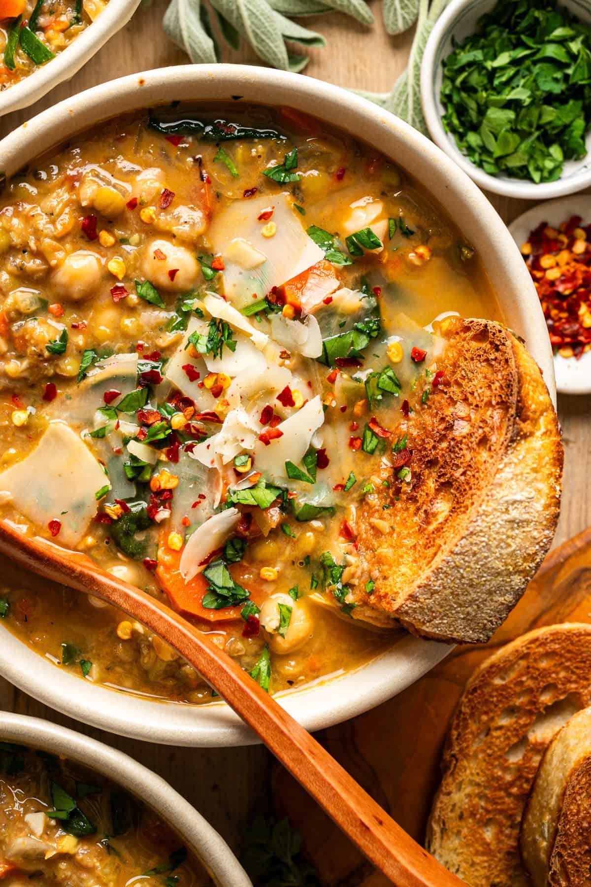 A bowl of hearty lentil soup with vegetables and greens, garnished with shaved cheese, chopped herbs, and red pepper flakes, served with a slice of toasted bread and a wooden spoon. Small bowls of herbs and chili flakes are nearby.