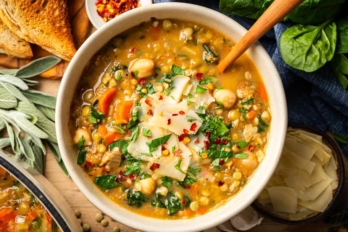 A bowl of vegetable and lentil soup topped with shaved cheese, chopped herbs, and red pepper flakes. Slices of toasted bread, fresh spinach, sage leaves, and a small bowl of cheese are nearby on a wooden surface.