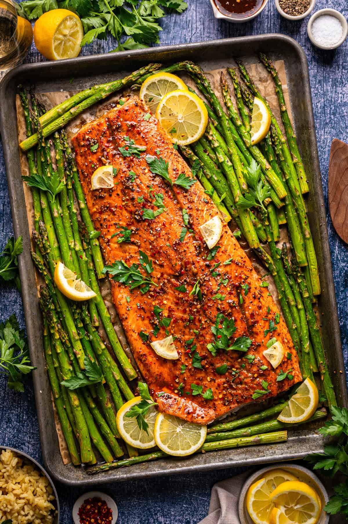 A baked salmon fillet garnished with lemon slices and parsley is surrounded by roasted asparagus spears on a baking sheet, with additional lemon wedges and fresh herbs nearby.